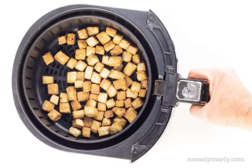 A hand holds the handle of an air fryer basket, full of a fresh batch of air fryer tofu.