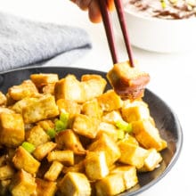 A hand holds chopsticks with a piece of air fryer tofu in it with dipping sauce on it. The full plate of tofu is behind it.