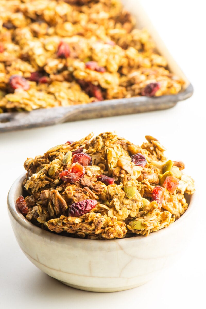 A bowl of protein granola sits in front of a pan with the rest of the granola, fresh from the oven.