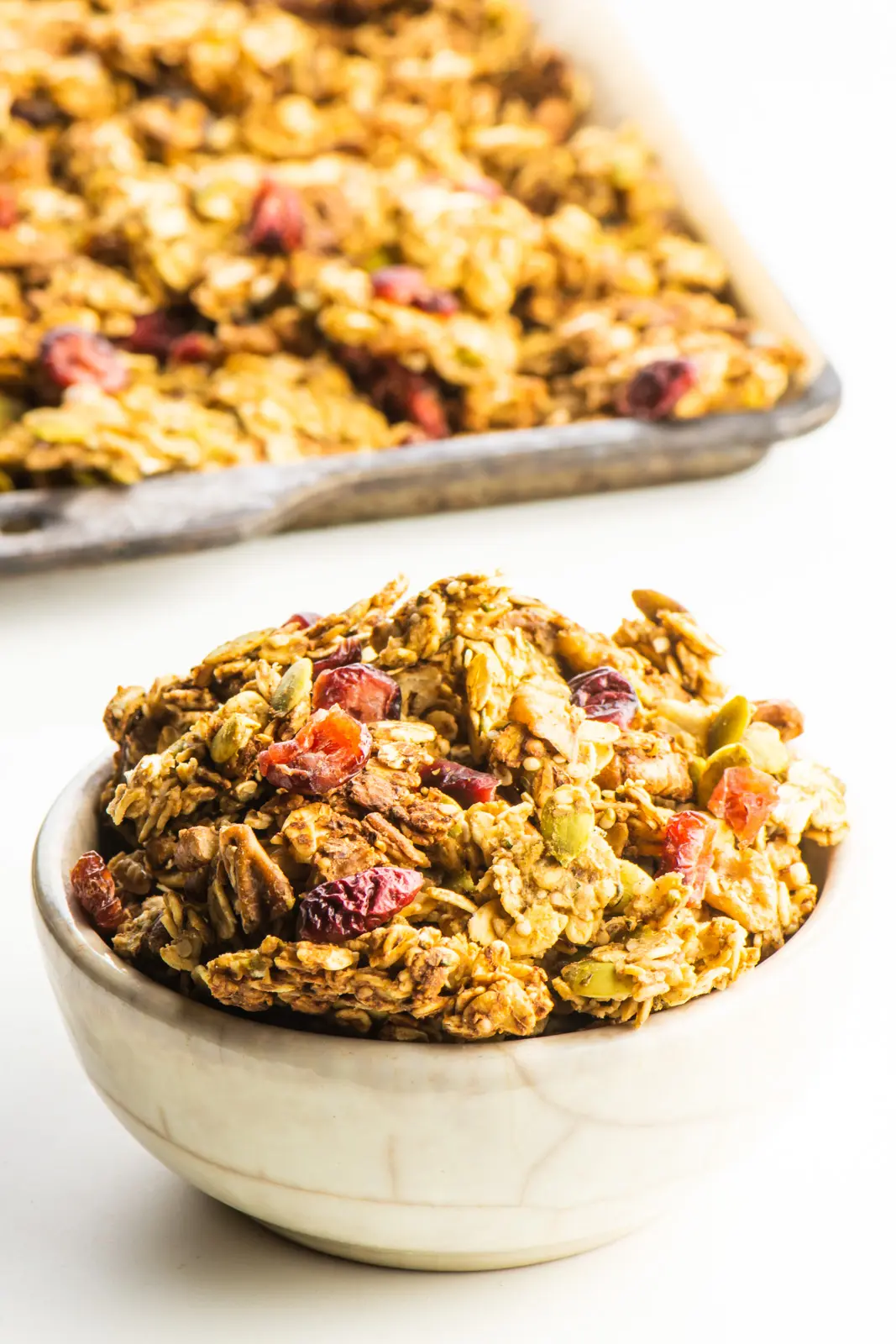 A bowl of protein granola sits in front of a pan with the rest of the granola, fresh from the oven.
