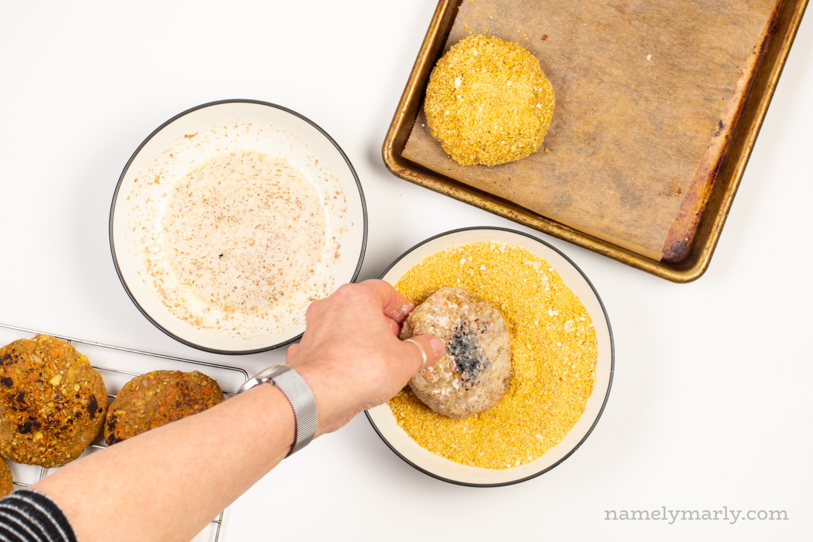 A hand is dipping a veggie patty in a bowl full of bread crumbs. There is a breaded patty on a baking sheet and another bowl for dipping.