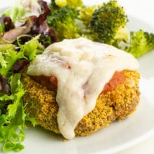 A vegan chicken parmesan patty with melted vegan cheese on a plate beside steamed broccoli and salad.
