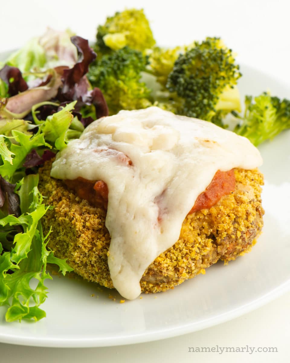 A vegan chicken parmesan patty on a plate with marinara sauce and melted vegan cheese. It sits beside a salad and steamed broccoli.