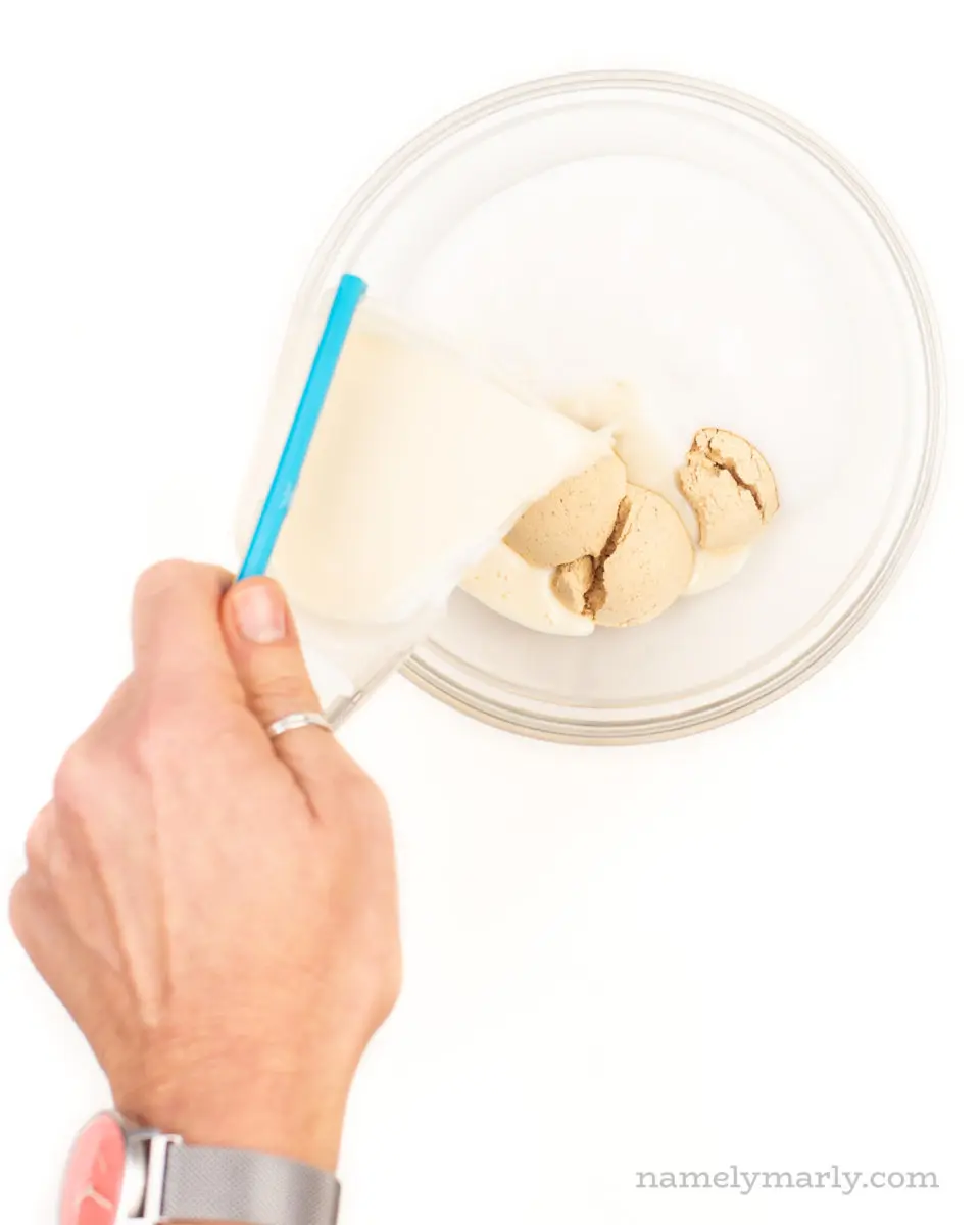A hand holds a measuring cup and pours plant-based milk over peanut butter powder.