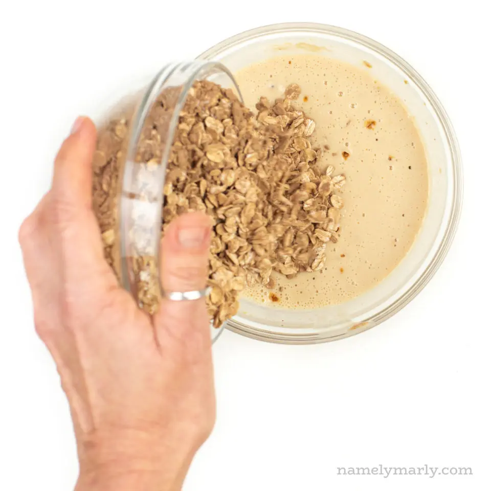 A hand holds a glass bowl and is pouring the contents of oatmeal into a plant-based milk mixture.