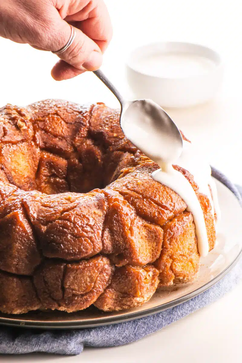 A hand holds a spoon pouring vanilla frosting over monkey bread.