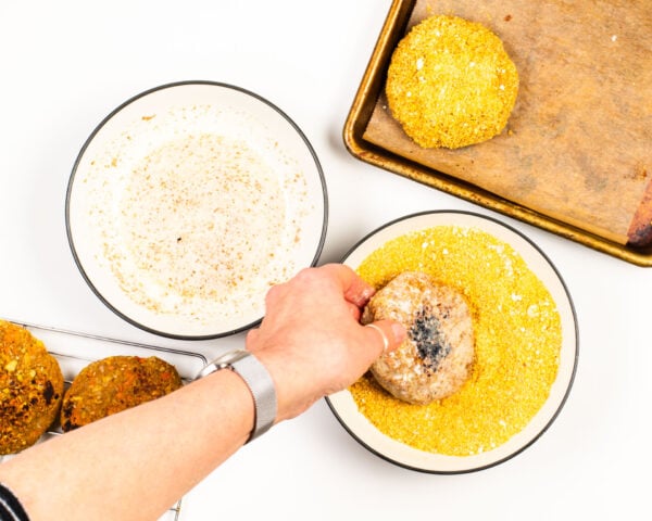 A hand is dipping a veggie patty in a bowl full of bread crumbs. There is a breaded patty on a baking sheet and another bowl for dipping.