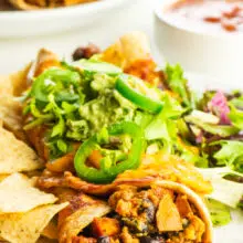 A vegan enchilada is topped with chopped lettuce and jalapeños with a bowl of salsa behind it.