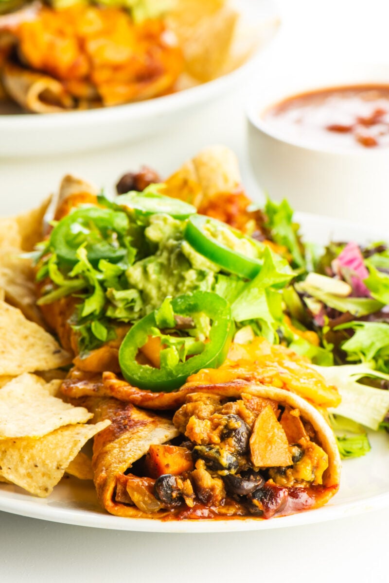 A vegan enchilada is topped with chopped lettuce and jalapeños with a bowl of salsa behind it.