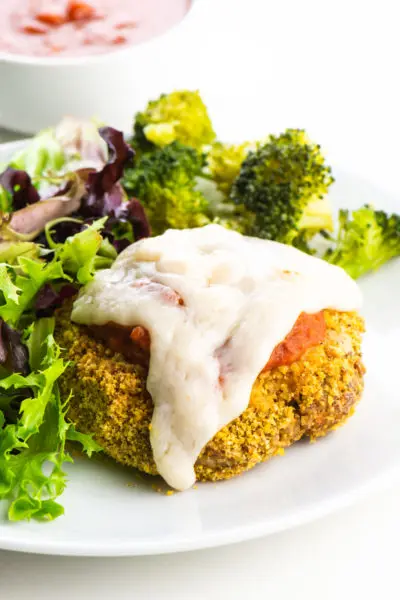 A vegan chicken parmesan patty on a plate beside steamed broccoli and salad.
