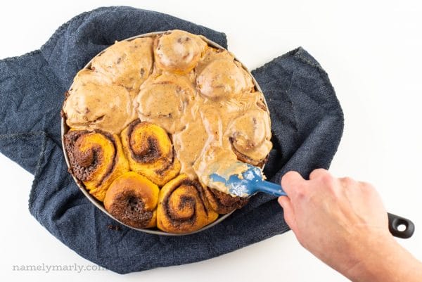 A hand holds a spatula spreading frosting over freshly baked cinnamon rolls.