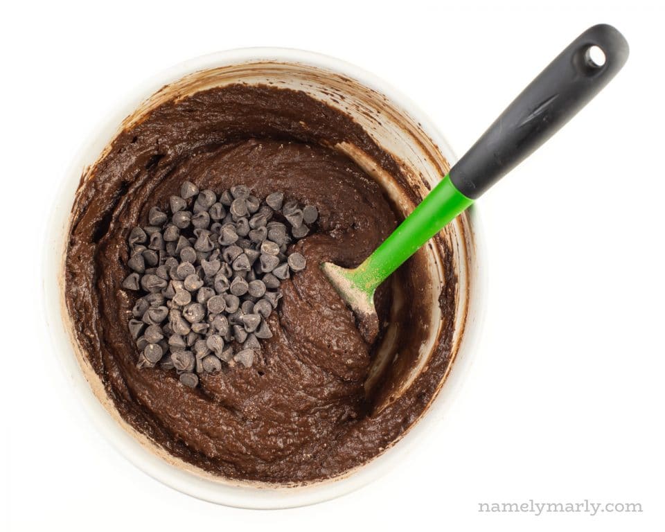 Chocolate batter is in a mixing bowl with chocolate chips. A green spatula is in the bowl.