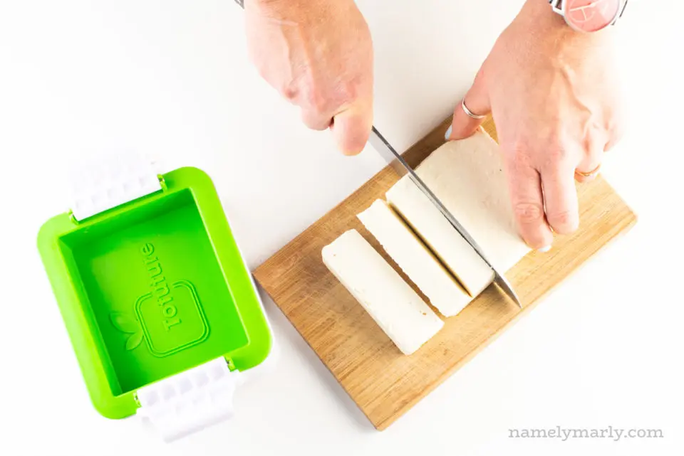 A hand holds a knife and is cutting tofu. It's sitting next to a tofu presser gadget.