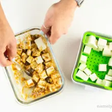 A hand uses a spoon to stir tofu with marinade. The rest of the cubed tofu sits in a green container.
