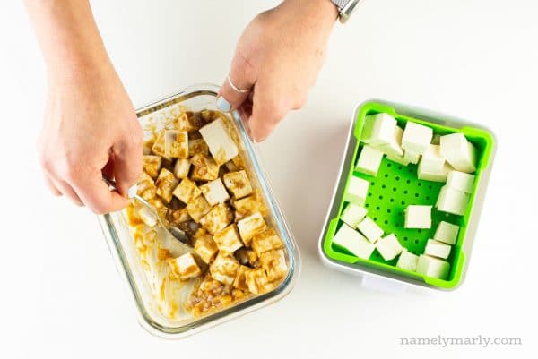A hand uses a spoon to stir tofu with marinade. The rest of the cubed tofu sits in a green container.