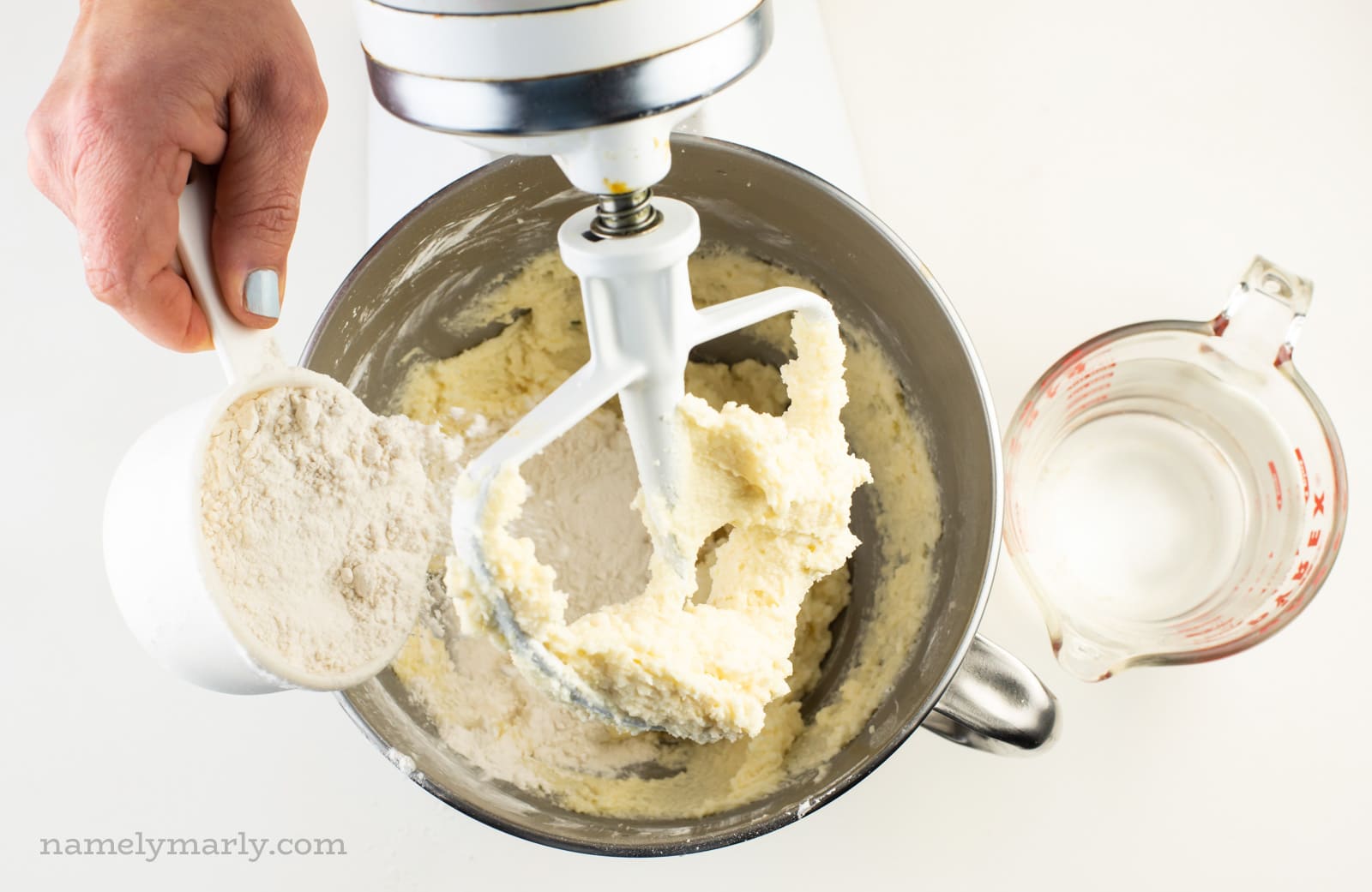 A hand pours flour into a mixing bowl. A pyrex measuring dish of water sits next to it.
