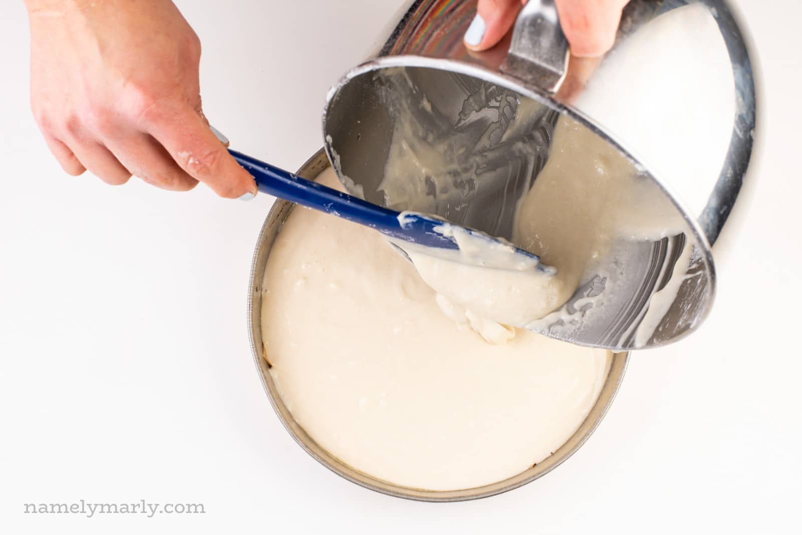A hand holds a spatula spooning cake batter from a mixing bowl to a cake pan.