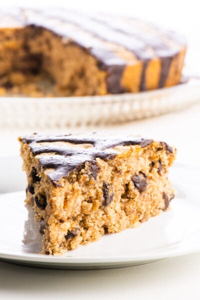 A slice of almond butter cake with chocolate drizzle on the top sits in front of the rest of the cake.
