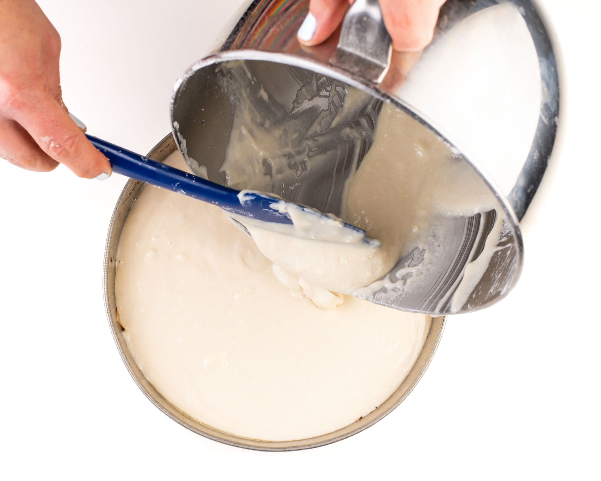 A hand holds a spatula spooning cake batter from a mixing bowl to a cake pan.