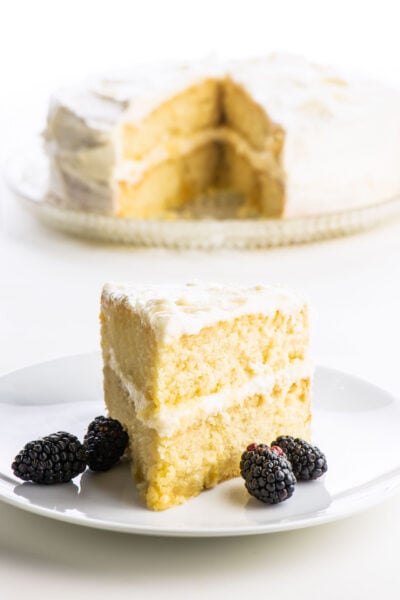 A slice of two-layer vegan vanilla cake with blackberries sitting next to it is in front of the rest of the cake.