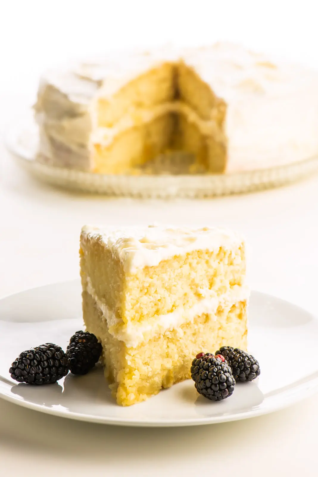 A slice of two-layer vegan vanilla cake with blackberries sitting next to it is in front of the rest of the cake.