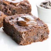 A slice of chocolate banana brownies with a dollop of melted chocolate over the top. A white bowl full of chocolate chips sits nearby.