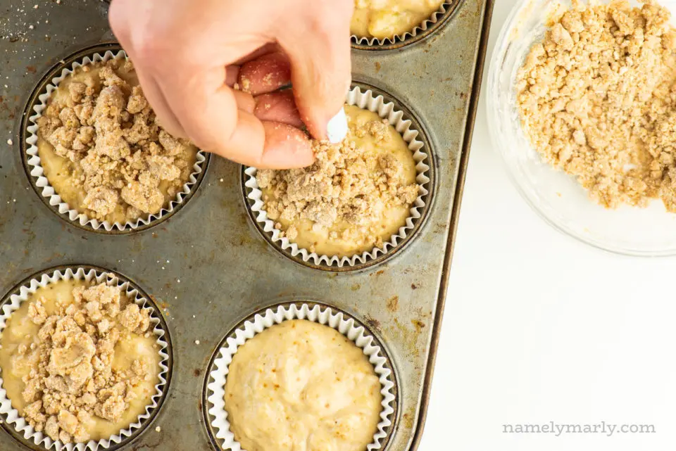 A hand is placing streusel crumbles on top of muffin batter in a muffin tin.