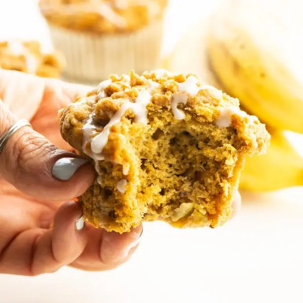 A hand holds a banana muffin with a bite taken out. More muffins and bananas are behind it.