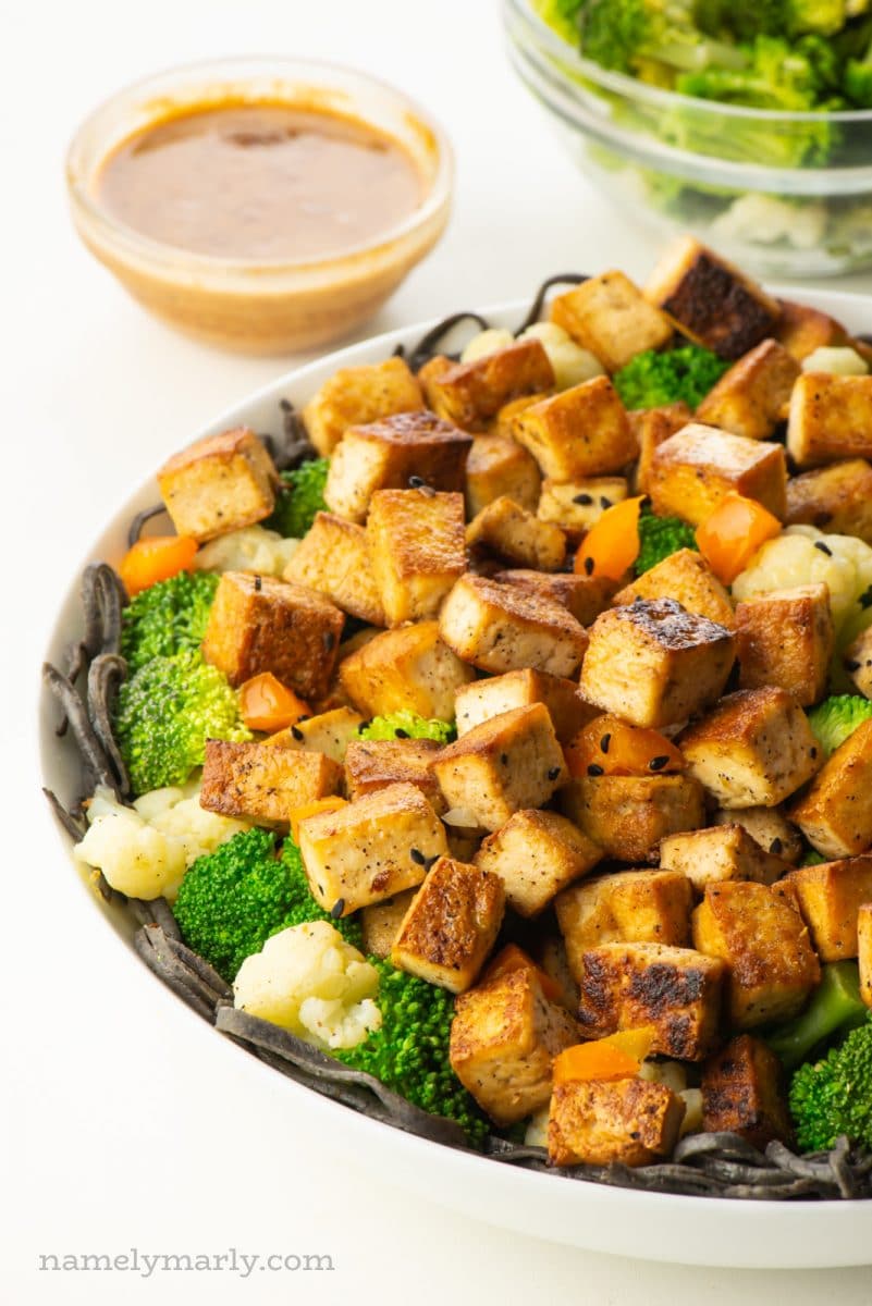 A bowl full of marinated tofu with steamed broccoli sits in front of a bowl of sauce and more broccoli.