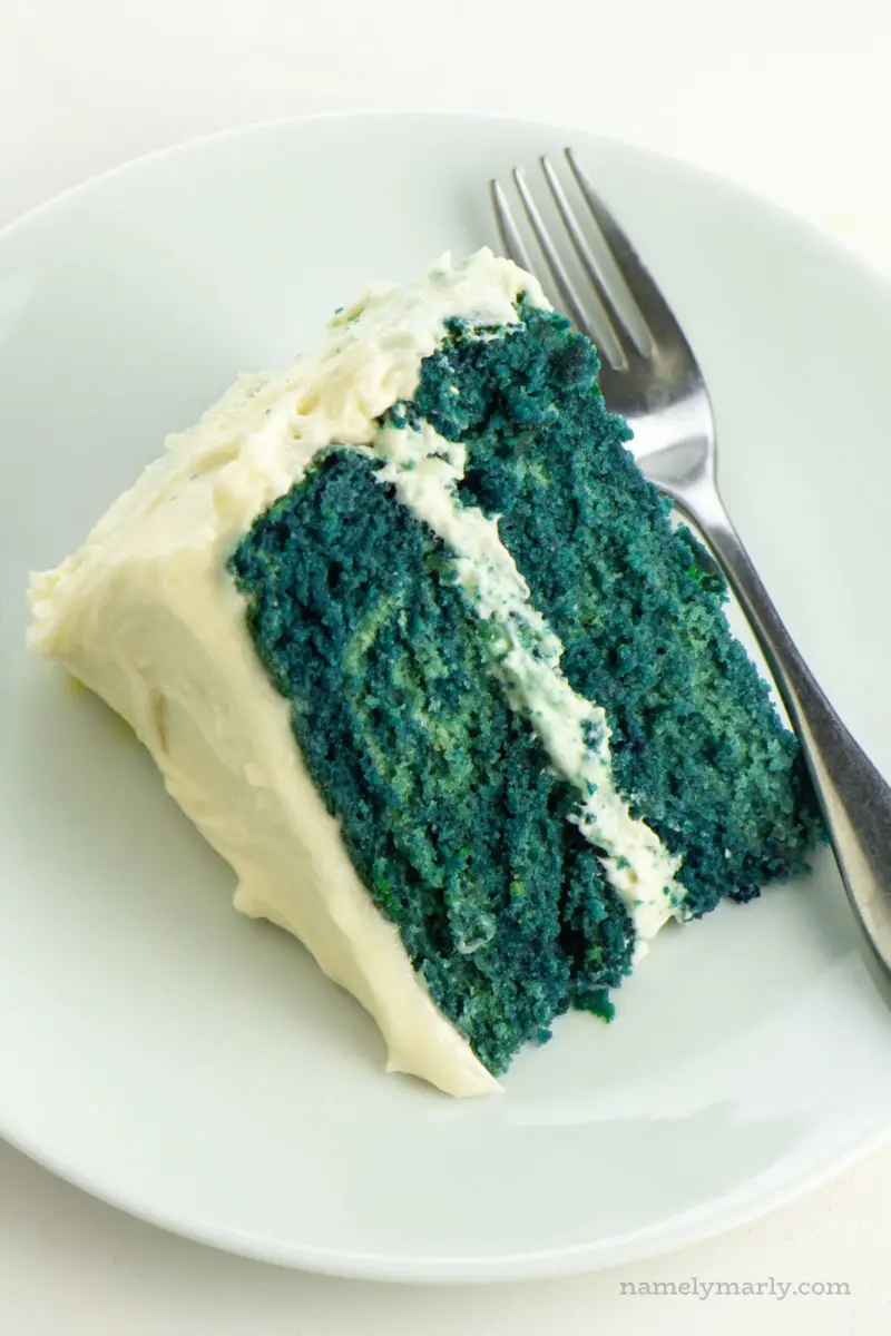 A slice of blue velvet cake lays on its side on a plate with a fork next to it.