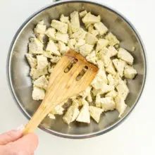 Tofu is in a skillet and a hand holds a spatula breaking the tofu into smaller pieces.