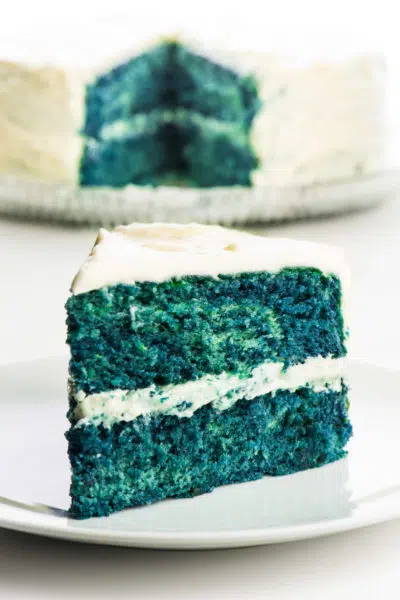 A slice of blue velvet cake on a plate in front of the rest of the cake.