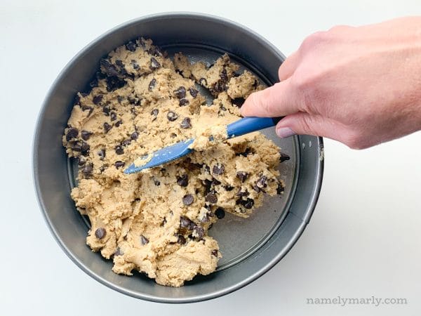 A hand uses a spatula to press cookie cake dough into a pan.