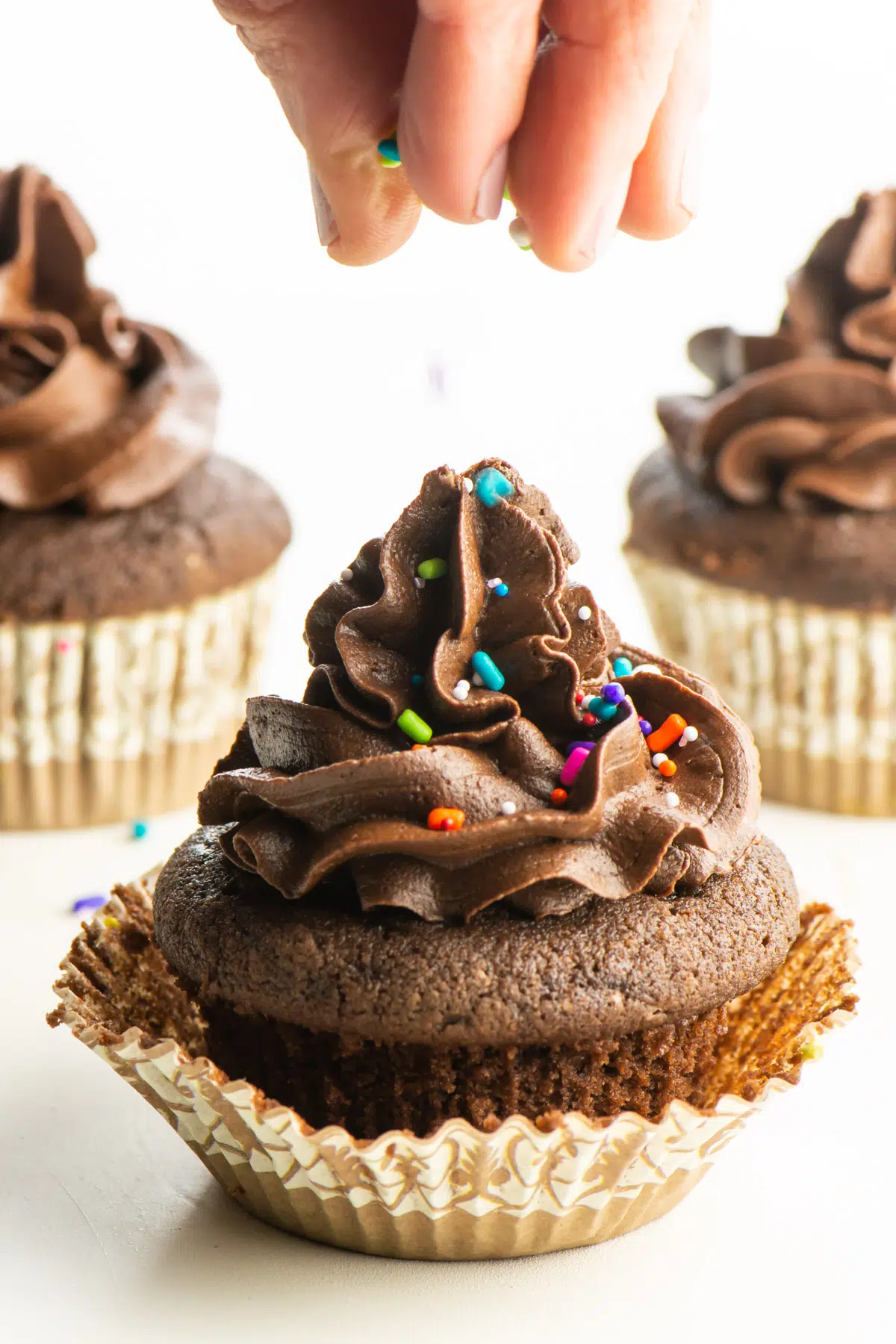 A hand drops sprinkles over a chocolate cupcake.