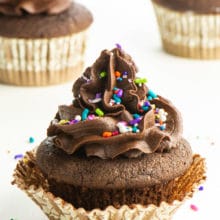 A cupcake topped with vegan chocolate frosting and sprinkles sits in front of 2 other cupcakes.