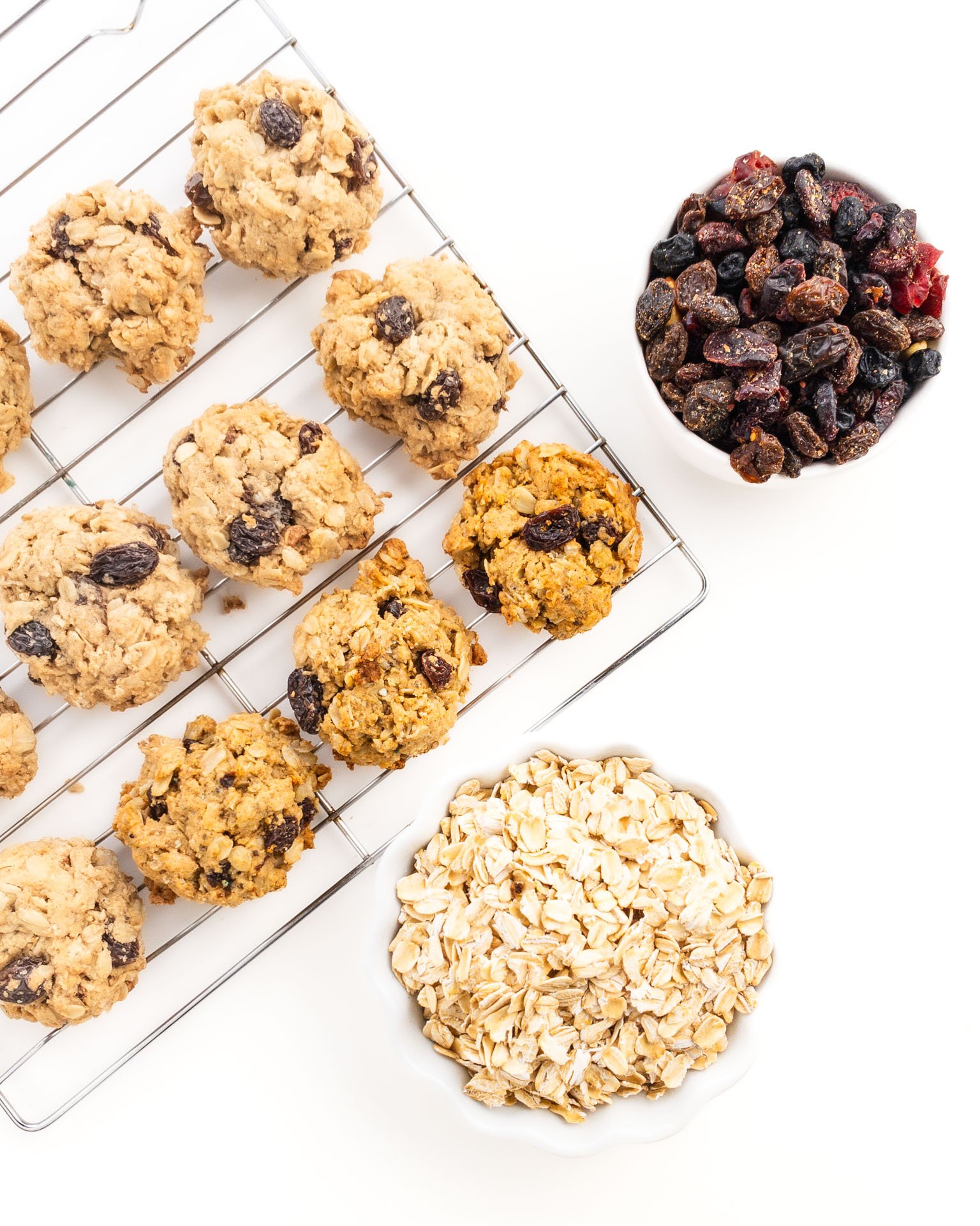 Cookies are on a baking rack next to oatmeal and a bowl of raisins.