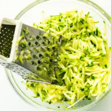A glass bowl contains shredded zucchini along with a metal shredder.
