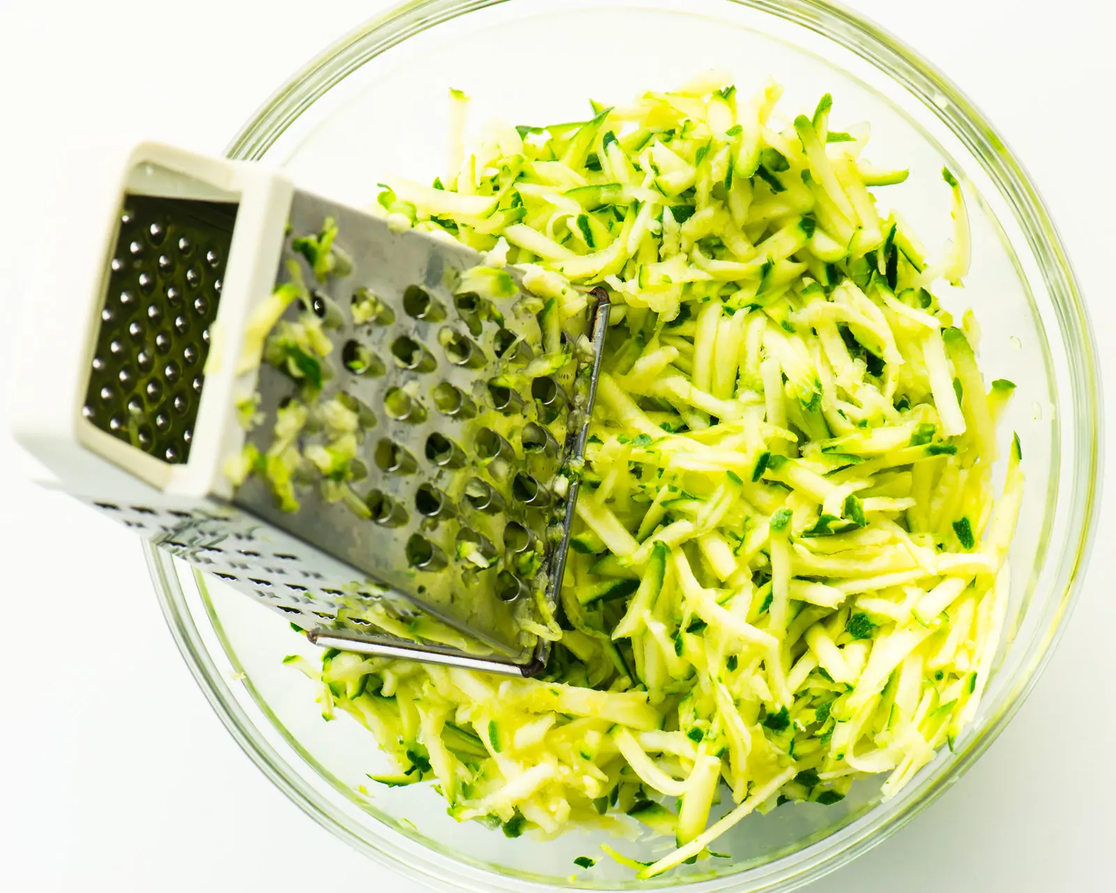 A glass bowl contains shredded zucchini along with a metal shredder.