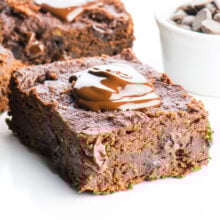 A slice of chocolate banana brownies with a dollop of melted chocolate over the top. A white bowl full of chocolate chips sits nearby.