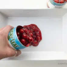 A hand pours a can of cherry pie filling into a white baking dish.