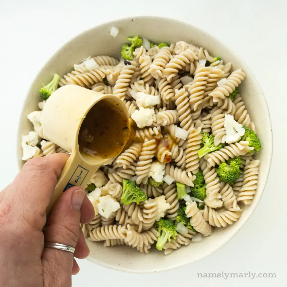A hand holds a measuring cup pouring dressing over pasta salad.
