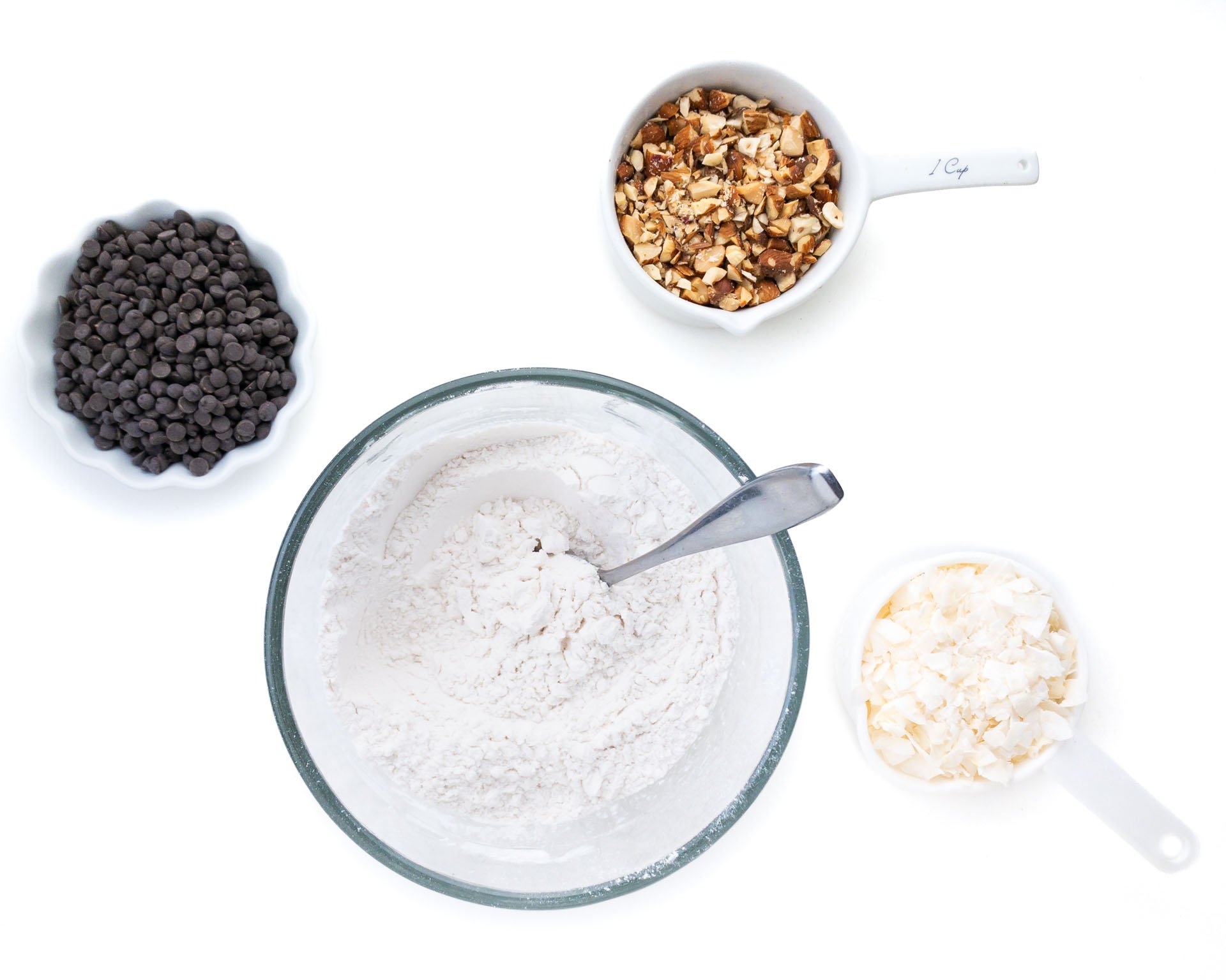 A glass bowl holds a flour mixture and is surrounded by bowls of coconut flakes, chocolate chips, and chopped almonds.