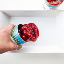 A hand pours a can of cherry pie filling into a white baking dish.