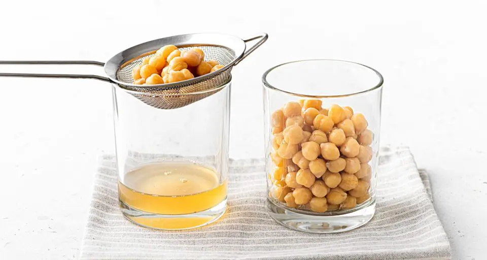 A glass holds chickpeas next to another glass with a strainer, with chickpea liquid in the bottom and chickpeas on the top of the strainer. Both glasses are sitting on a folded kitchen towel.