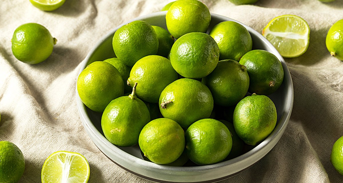A bowl full of key limes sits on a table lined with cloth. There are several limes around the bowl, some of them cut open to reveal the flesh of the fruit.