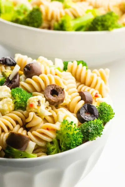 A bowl of pasta salad featuring black olives and broccoli sits in front of another bowl.