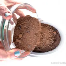 Two hands hold a glass bowl pouring chocolate crumbles over a cake batter.