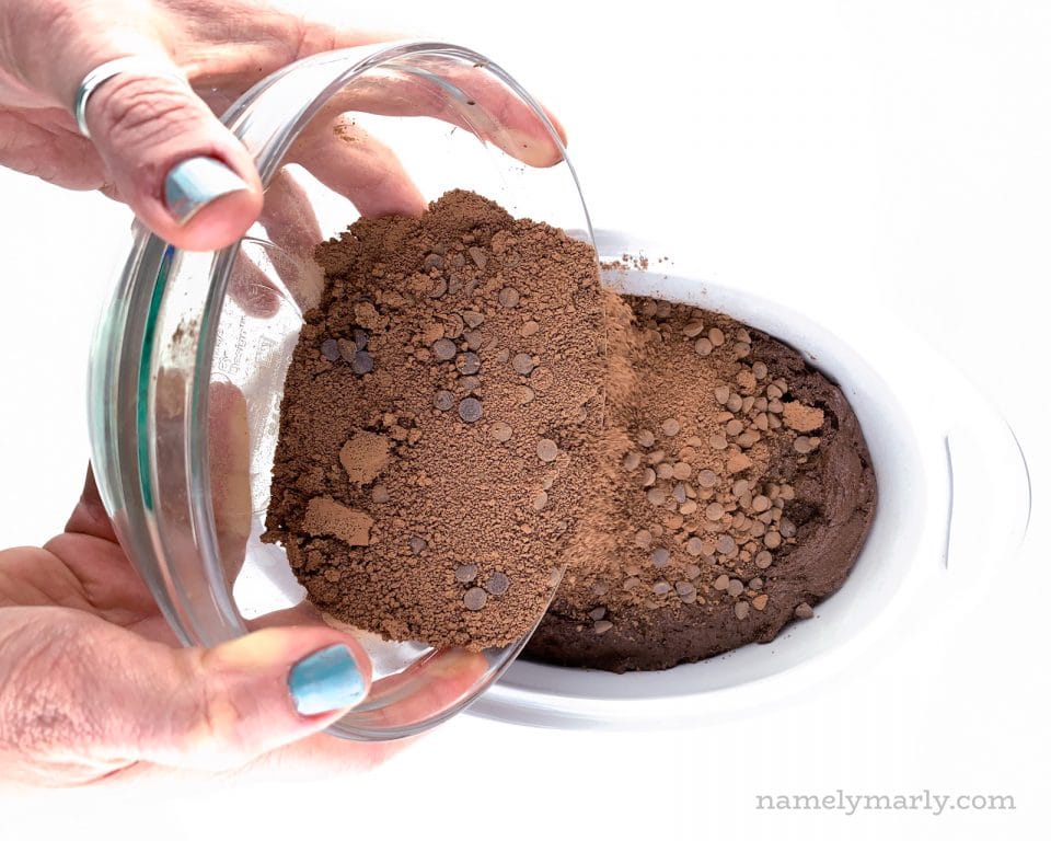 Two hands hold a glass bowl pouring chocolate crumbles over a cake batter.