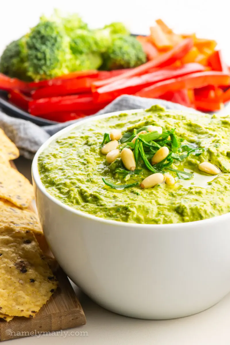 A bowl of green hummus sits next to chopped veggies and chips.