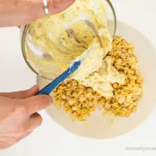 A hand holds a glass bowl over mashed chickpeas and uses a spatula in the other hand to spread it over the chickpeas.